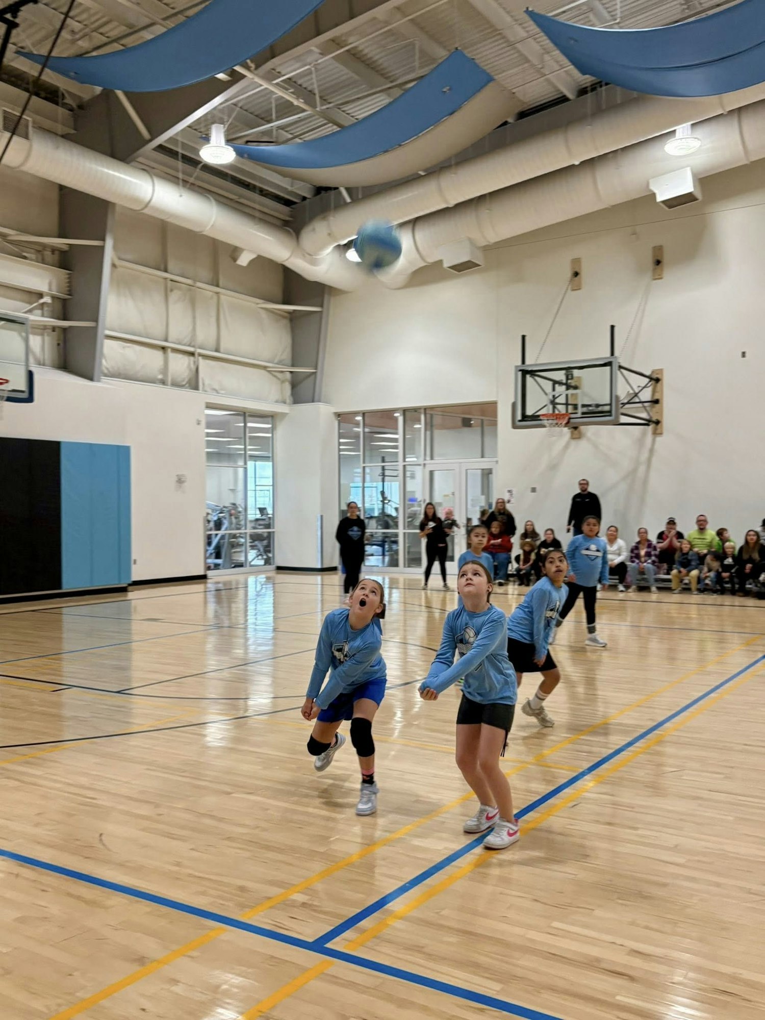 A youth sports event in a gym, with girls in light blue shirts playing and spectators watching from the sidelines.