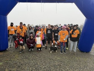 A group of people in orange shirts gathered under a blue archway, likely at a fun run or charity event on a rainy day.