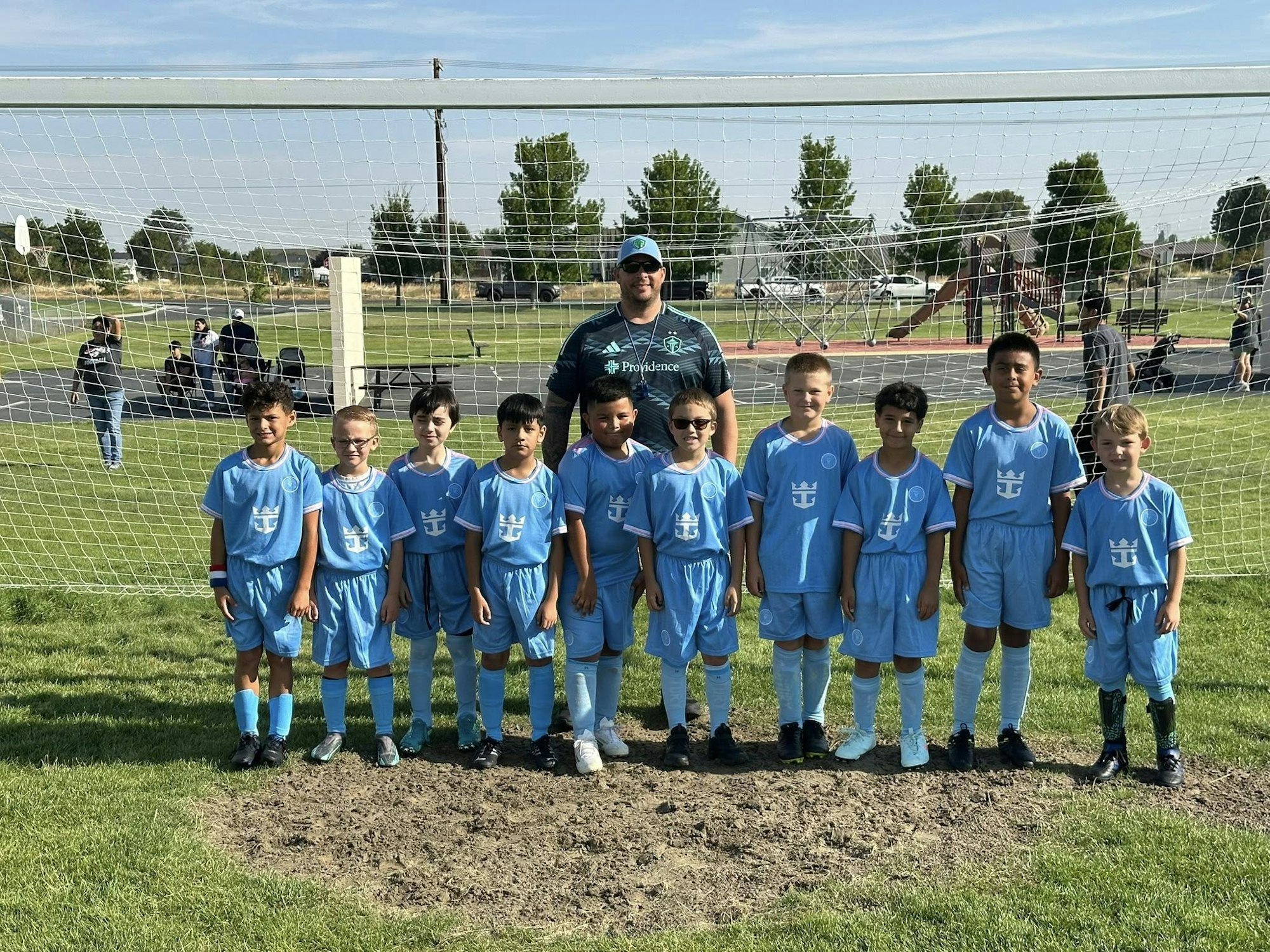 A youth soccer team in blue uniforms poses with their coach in front of a goal on a sunny day at a park.