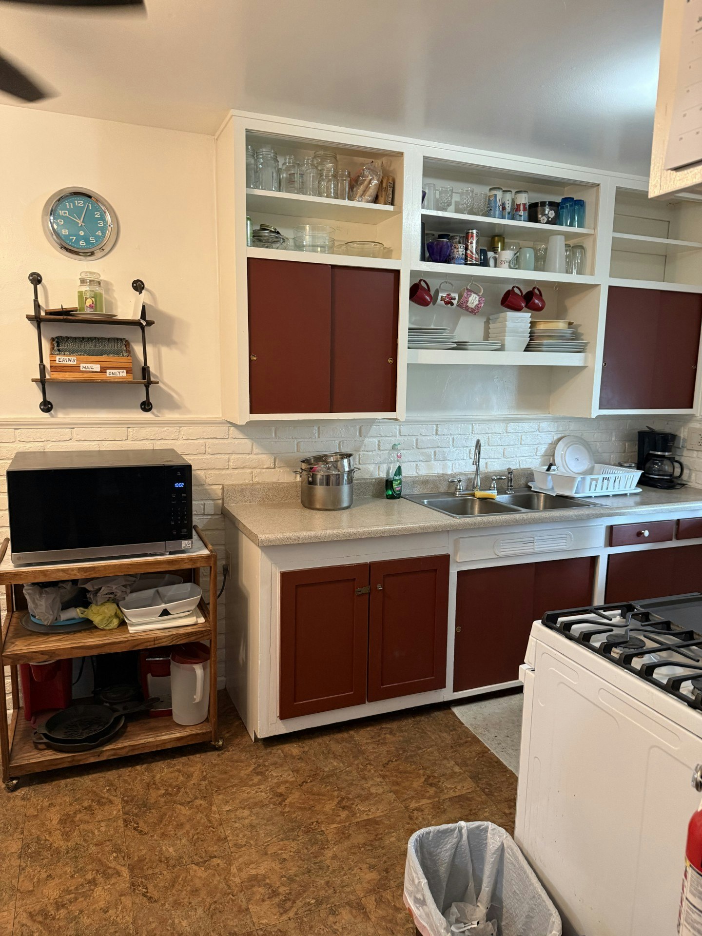 A kitchen with white cabinets, red doors, a blue clock, dishes, a microwave, a stove, and a trash can.