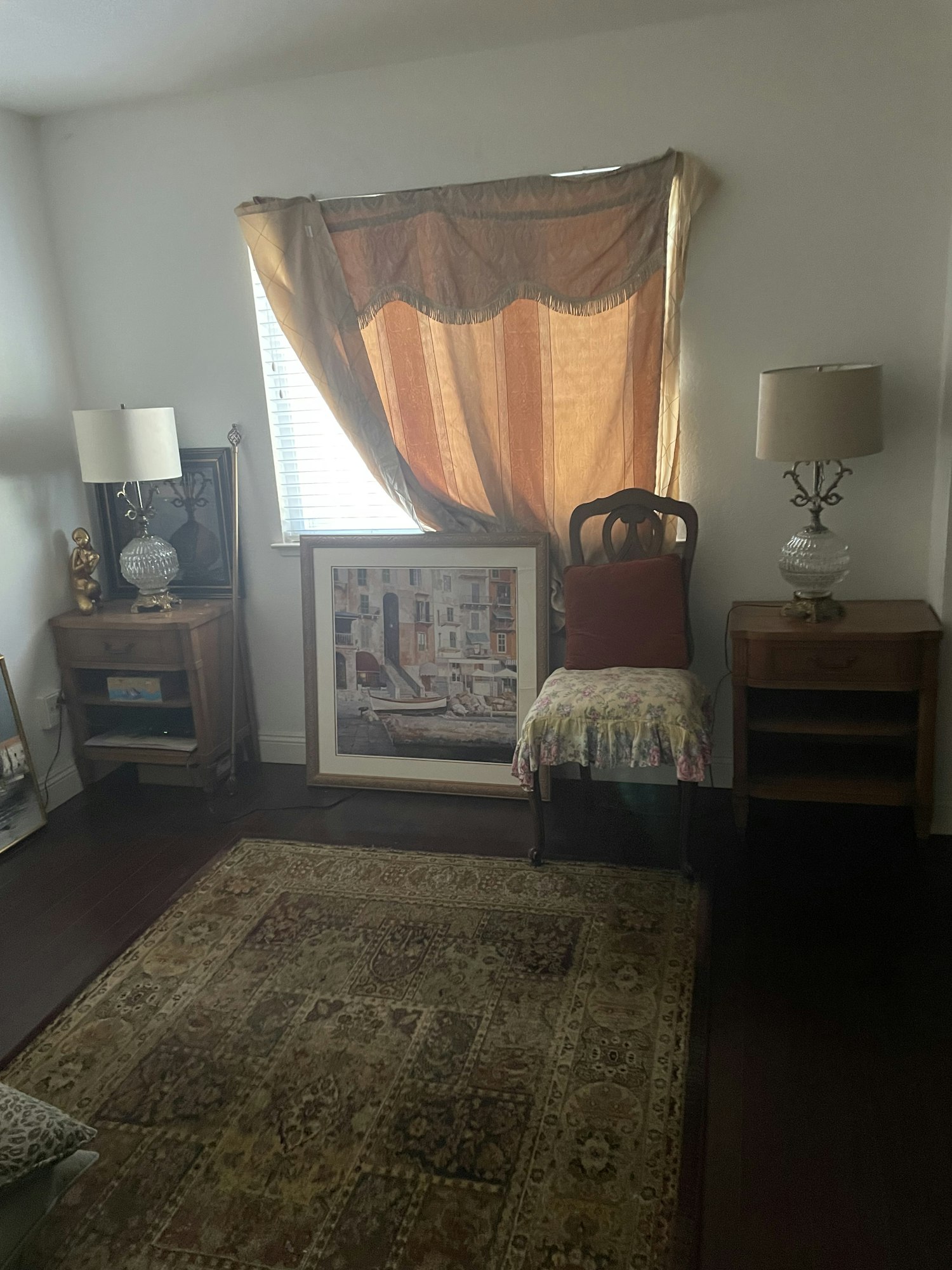 A cozy room with a decorative rug, framed artwork, wooden furniture, lamps, and a draped window.