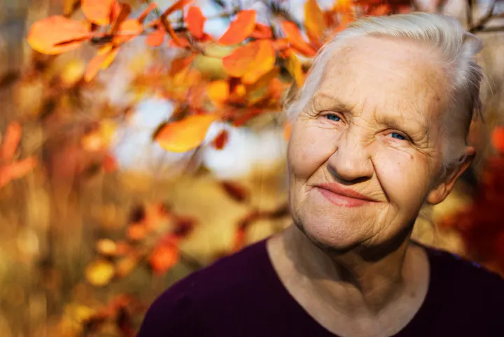 An elderly woman smiling, with sunlit autumn leaves in the background.