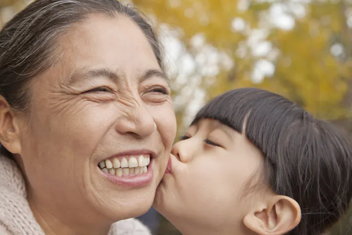 A woman smiles joyfully as a young girl kisses her cheek, surrounded by a warm, autumn atmosphere.