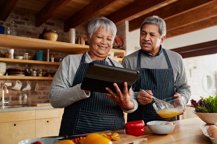 Two people in aprons cooking together in a kitchen, one holding a tablet and the other mixing ingredients.