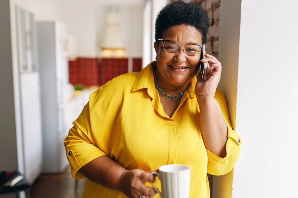 Woman in yellow shirt holding a cup and talking on the phone, smiling.