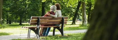 Two people sitting on a park bench surrounded by green trees.