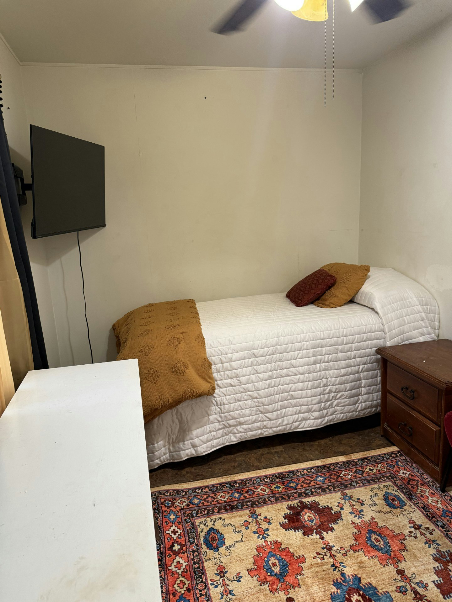 A small room with a single bed, colorful rug, TV on the wall, dresser, and side table.