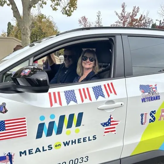 A woman and a man are smiling from inside a decorated car for Meals on Wheels, featuring patriotic symbols and flags.