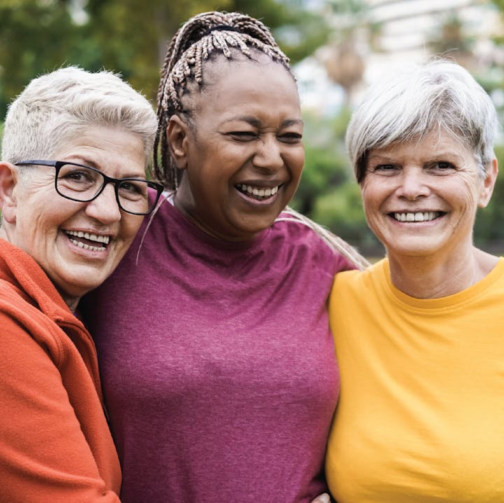 Three smiling women embracing, with a background of blurred greenery.