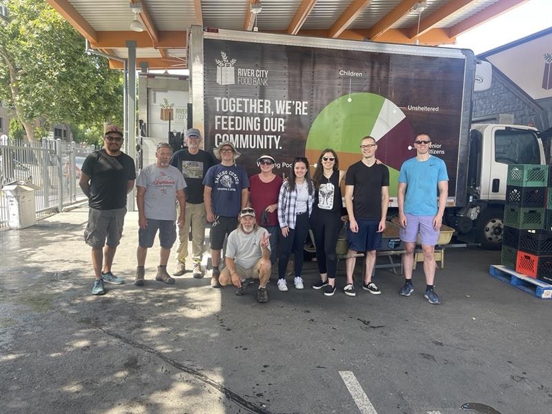 A group of people posing in front of a River City Food Bank truck with a community support message and chart.
