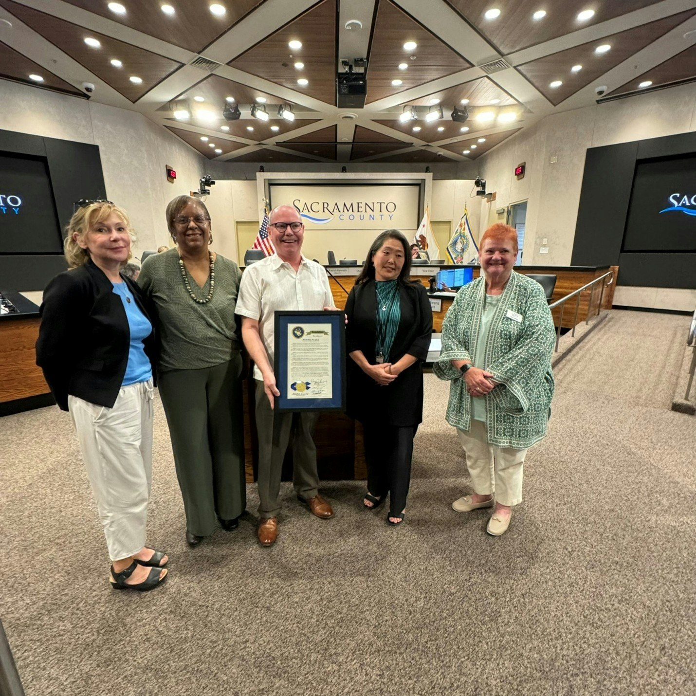 A group of five individuals stands together in a Sacramento County meeting room, holding a framed document.