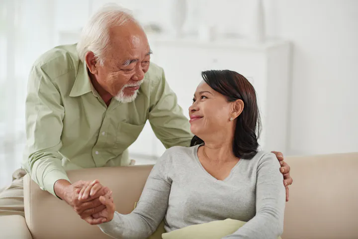 An older couple shares a warm moment, smiling and holding hands, in a cozy, bright living space.