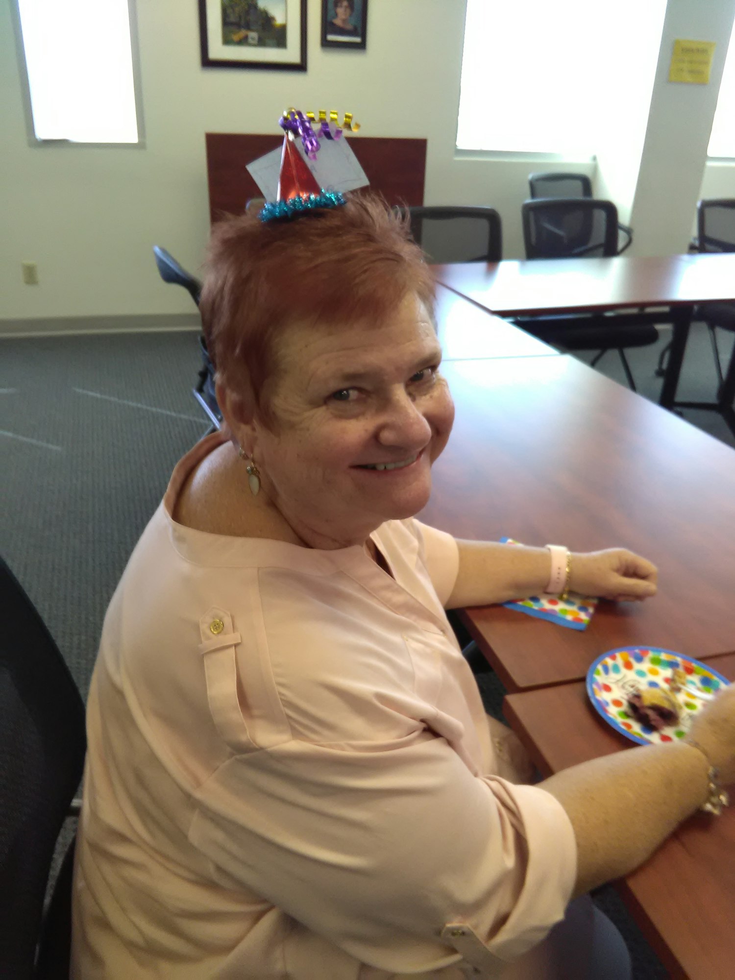 A smiling woman with short red hair wears a festive party hat while enjoying a treat at a table.