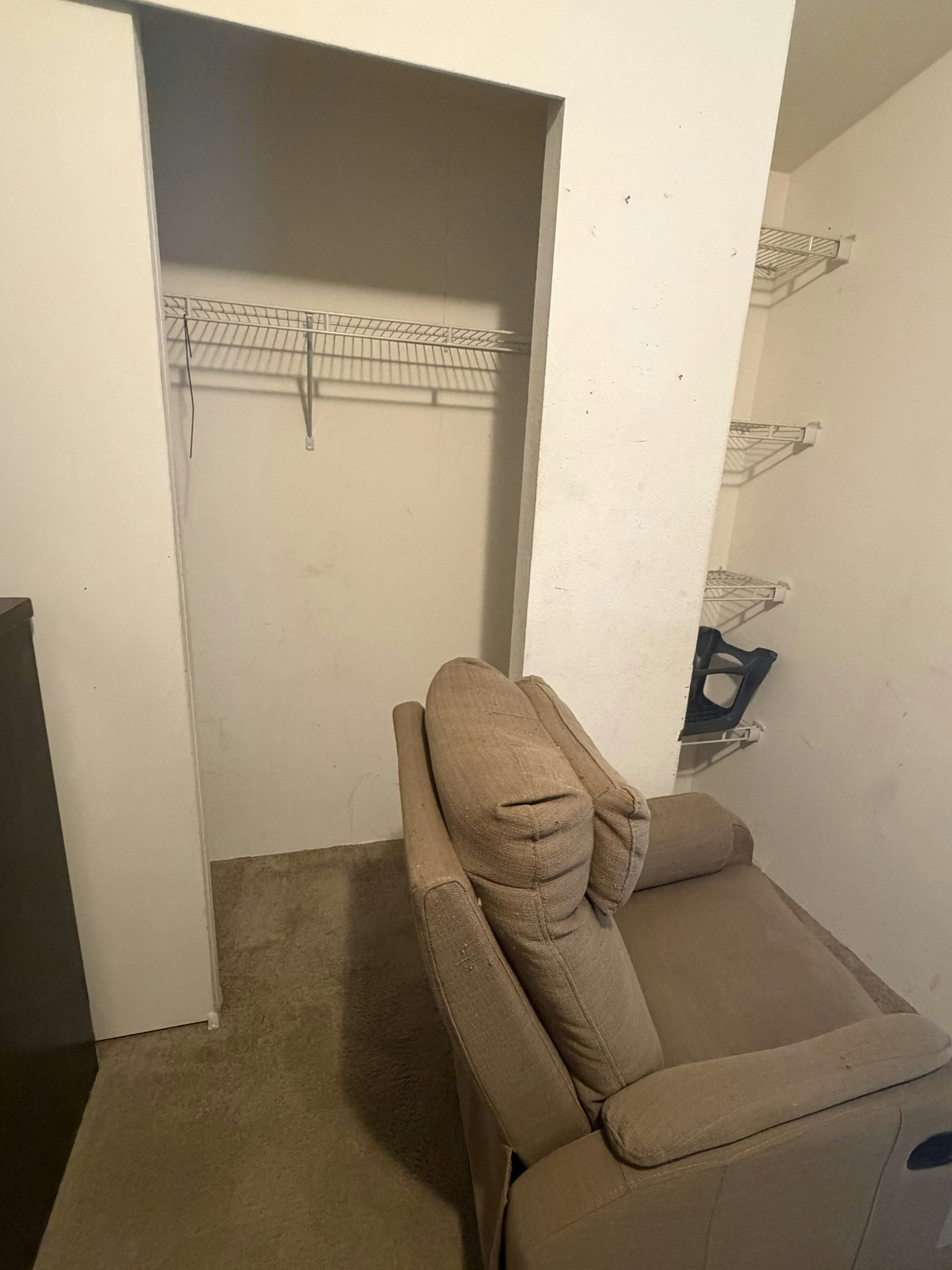 An empty closet space with a wire shelf, a beige chair beside it, and carpeted flooring.