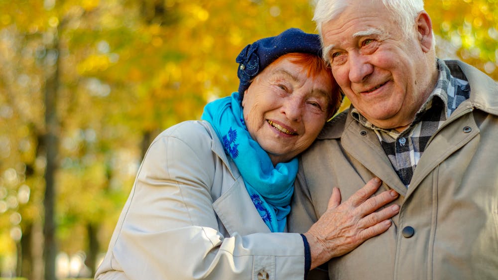 An elderly couple smiling and embracing each other in a park with autumn leaves in the background.