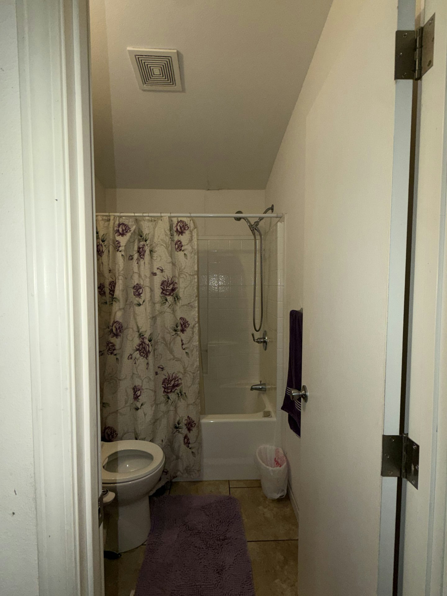 A bathroom featuring a shower with floral curtain, bathtub, toilet, and purple towel and mat.