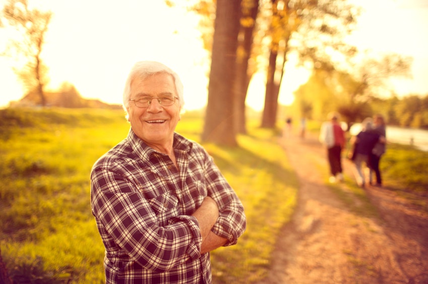 Older man smiling in a plaid shirt, standing on a sunny path with blurred people in the background.
