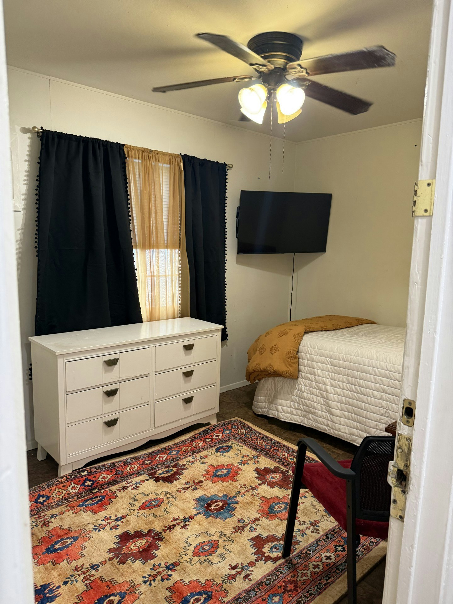 The image shows a cozy bedroom with a bed, chest of drawers, TV, chair, patterned rug, and curtains.