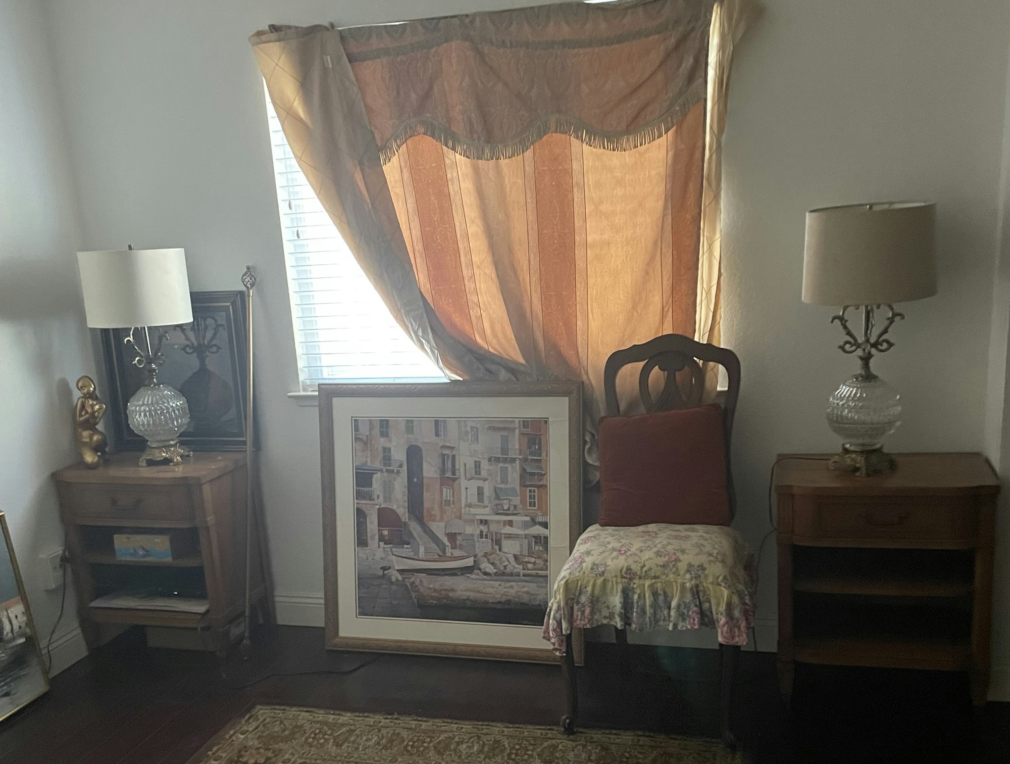 Room with a chair, two lamps on nightstands, a large framed painting, curtain-draped window, and decorative statue.