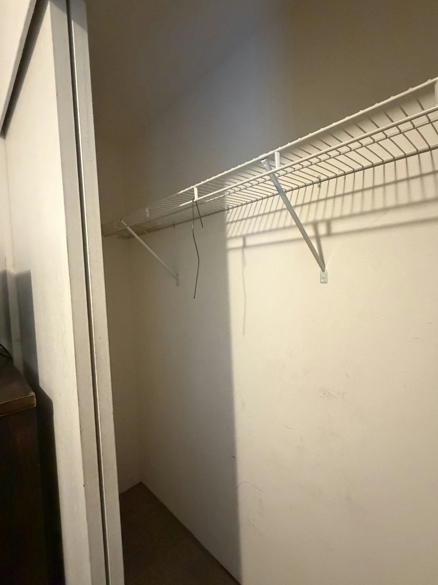 A small, empty closet with a wire shelf and minimal lighting, featuring plain walls and a shadow.