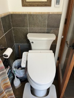 A bathroom with a toilet, toilet paper holder, trash can, and cleaning supplies.