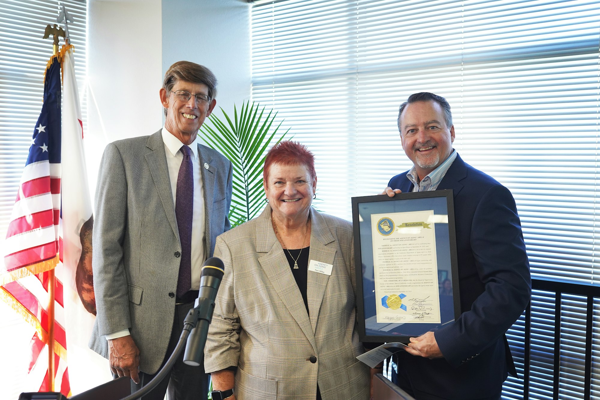 Three individuals are posing with a framed certificate in an office, with a California flag and a microphone in the foreground.