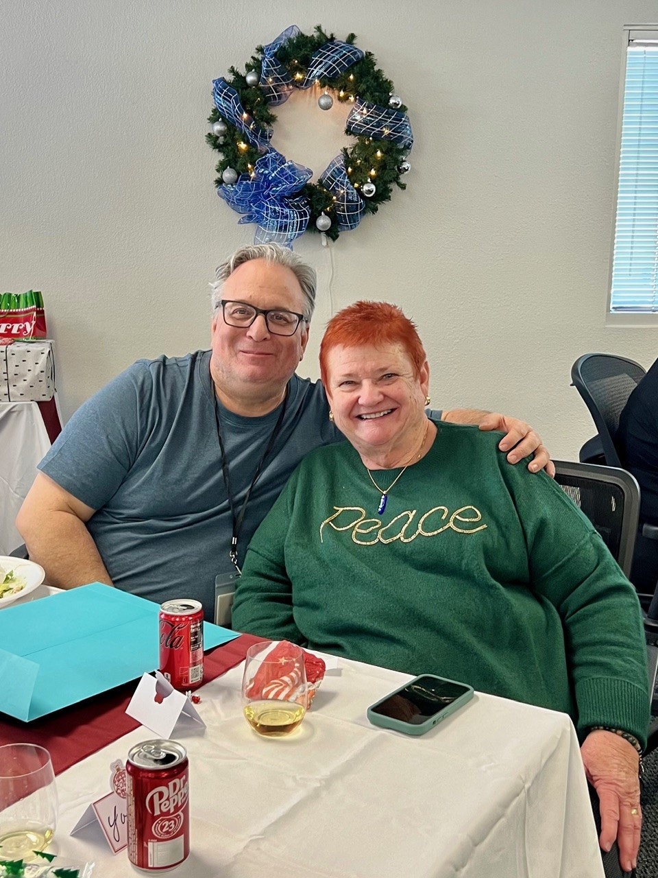 A man and woman sit together, smiling at a festive gathering with a holiday wreath in the background and drinks on the table.