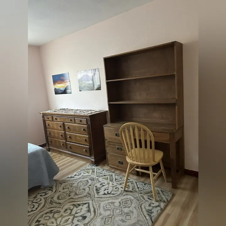 The image shows a room with wooden furniture, including a dresser, a bookshelf, a chair, and wall art.