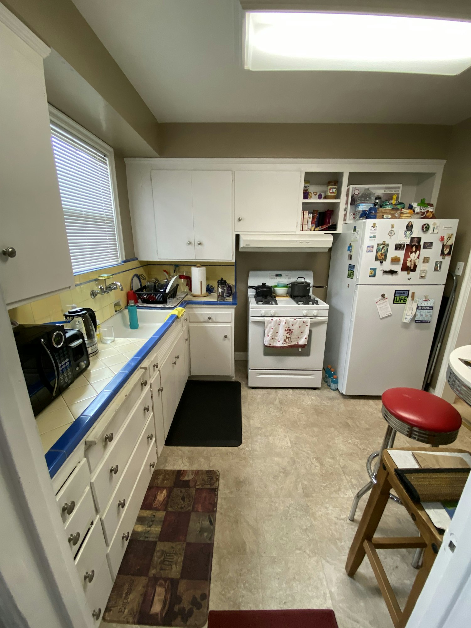 A small kitchen with white cabinets, fridge with magnets, stove, coffee maker, and counter with barstools.