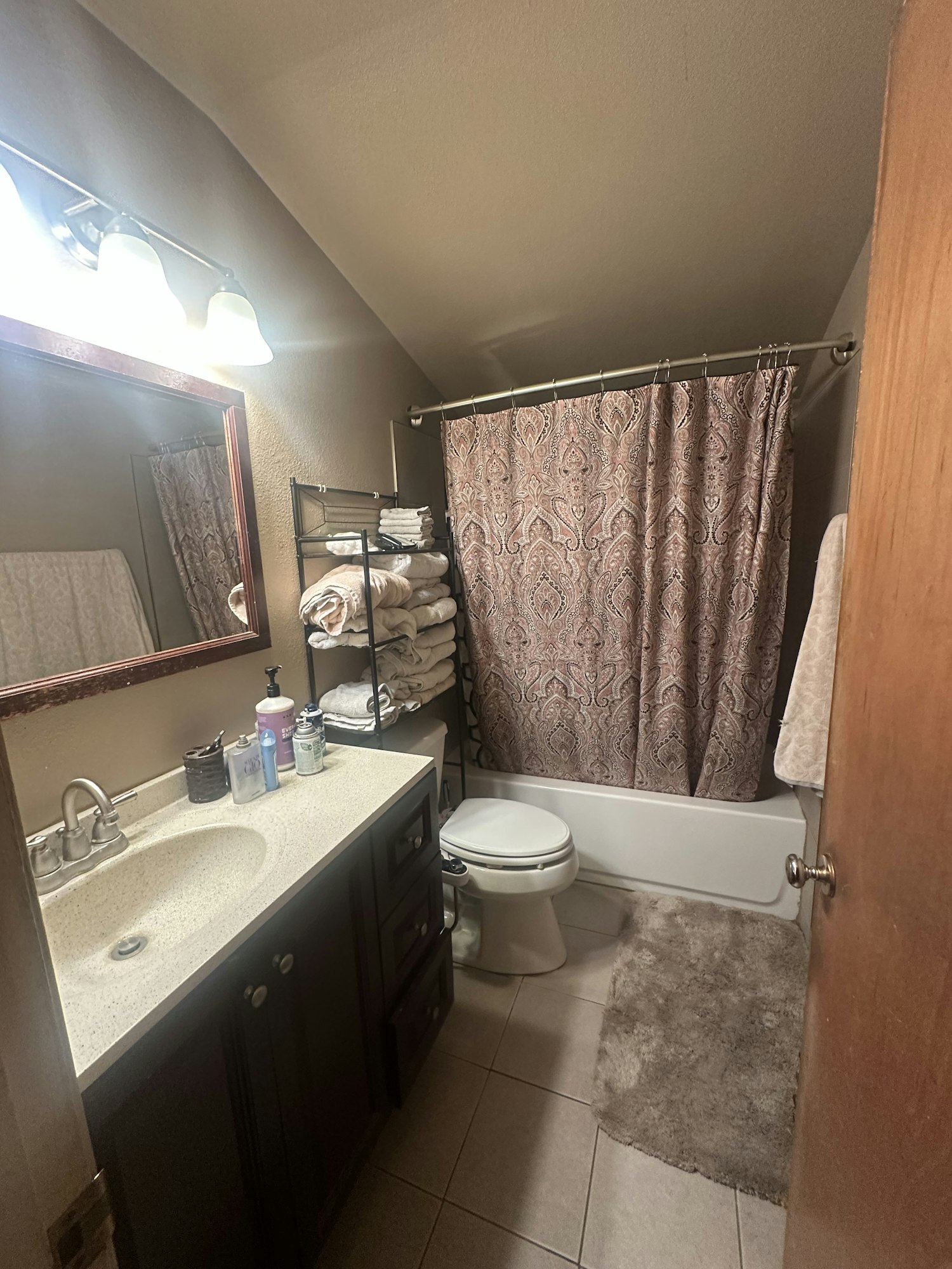 A bathroom with a shower, toilet, sink, mirror, and shelf with towels and toiletries.