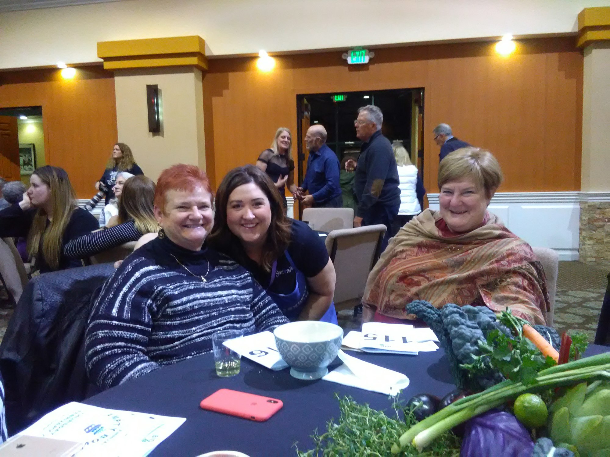 Three women are sitting together at a table, smiling, with a colorful assortment of vegetables in the foreground.