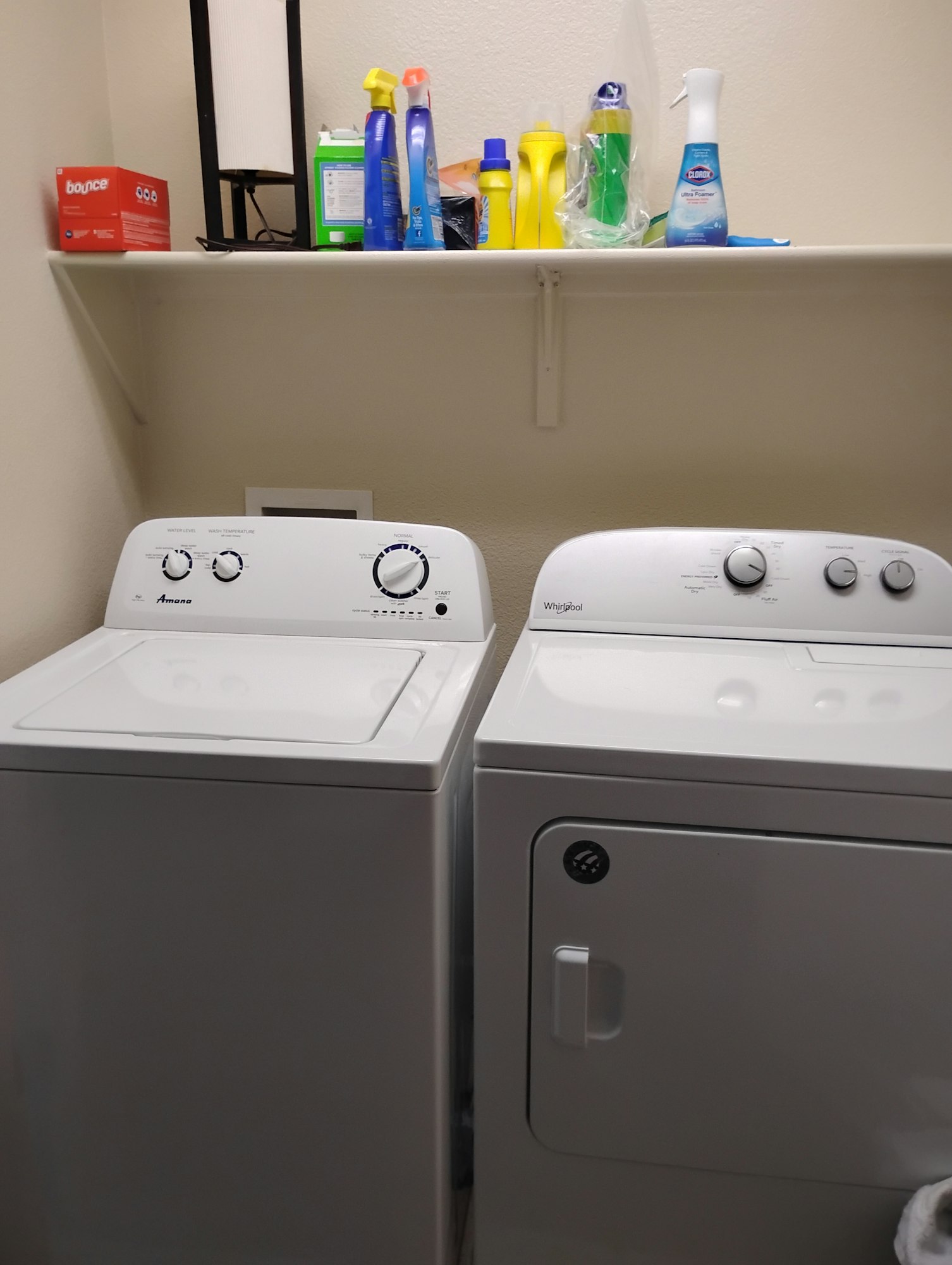 The image shows a stacked washer and dryer with shelf above holding various cleaning supplies.