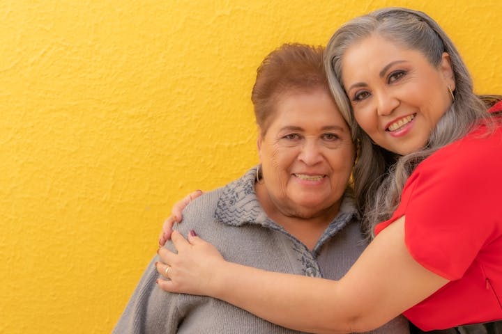 Two smiling women embracing in front of a yellow wall.