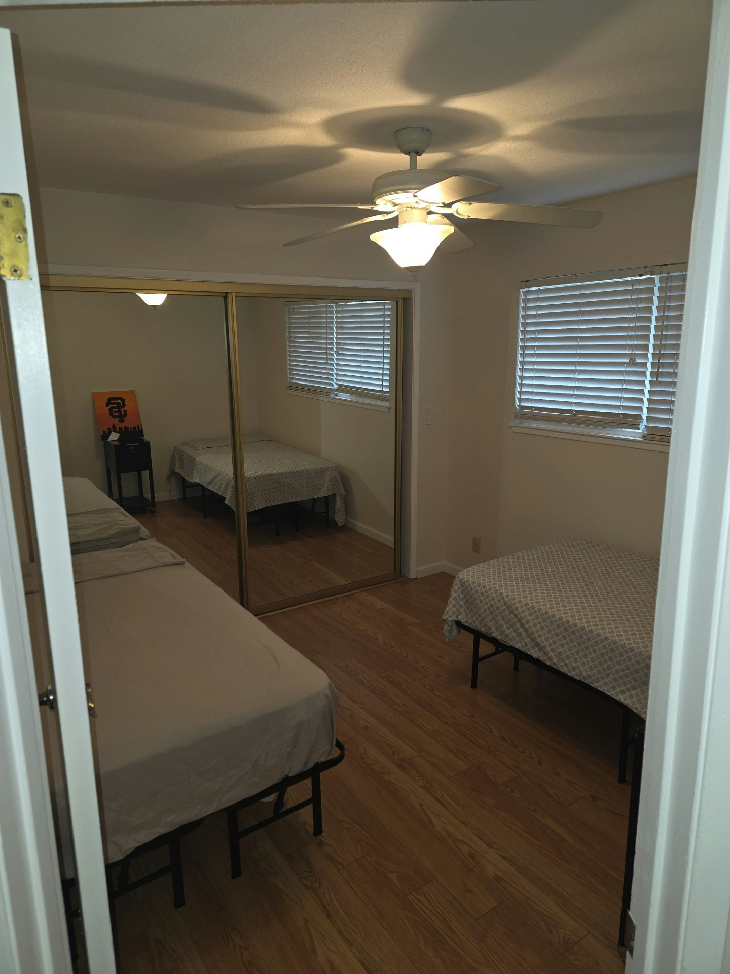 A tidy bedroom featuring two beds, a ceiling fan, and a large mirrored closet, with beige tones and wooden flooring.