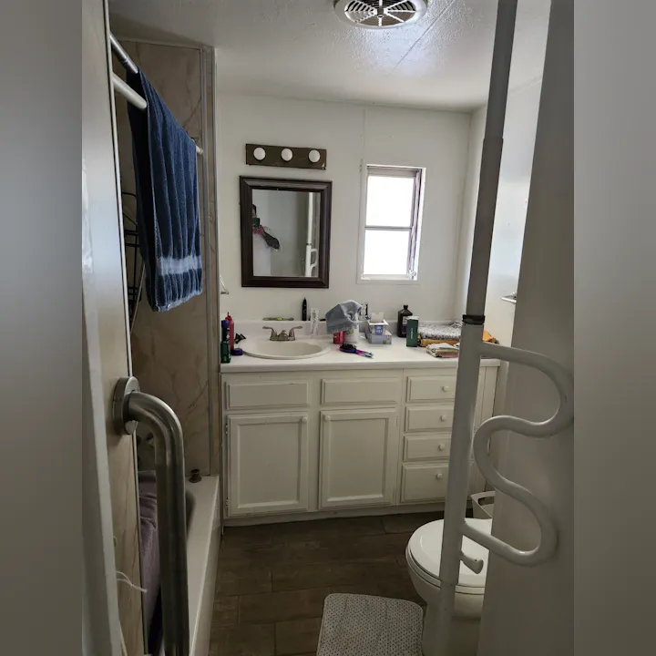 A small bathroom featuring a sink, mirror, toilet, shower, and towel rack, with various toiletries on the counter.