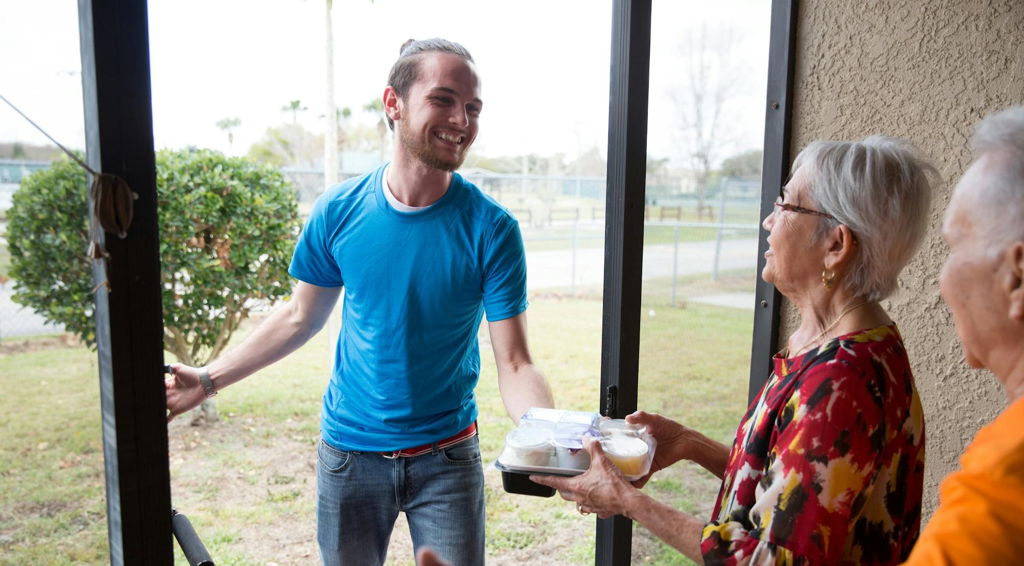 A man delivers food to elderly people at a doorway, all are smiling.