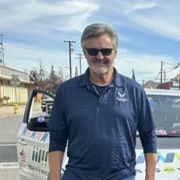 A man wearing sunglasses and a blue polo shirt stands in front of a vehicle decorated with various stickers.
