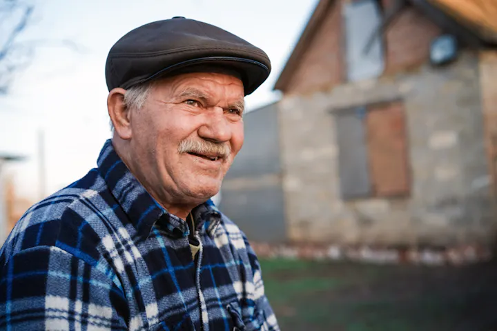 An older man with a smile wears a plaid shirt and a flat cap, sitting in front of a rustic house.