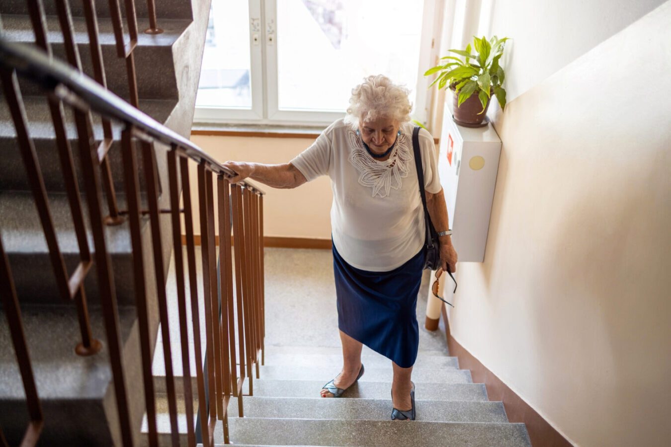An elderly woman is climbing stairs while holding onto the railing and wearing a light-colored blouse and dark skirt.