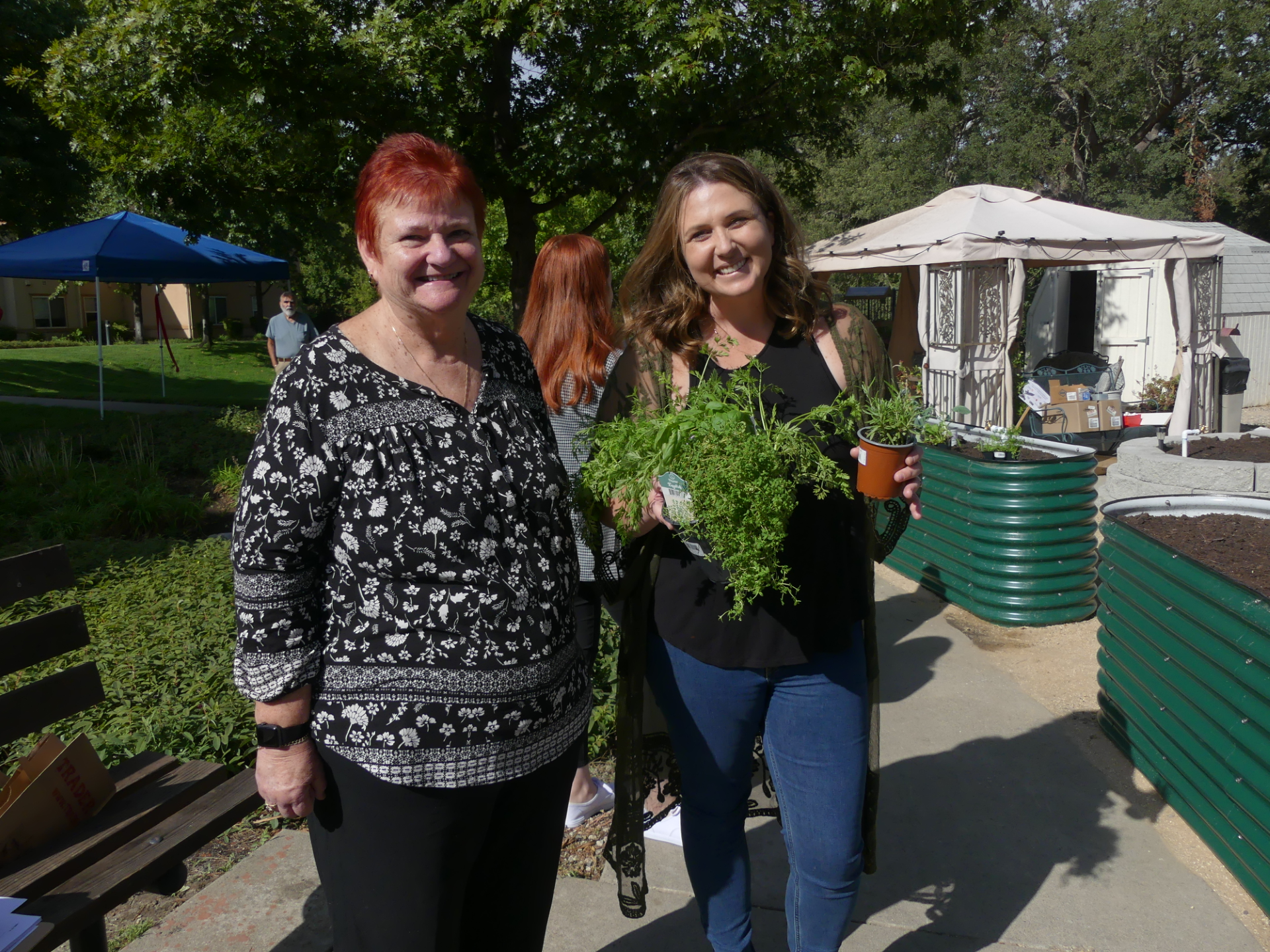 Two women smile outdoors, one holding potted herbs, with greenery and a tent in the background.