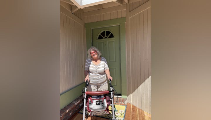 A smiling older woman with red glasses stands with a walker at a doorway with a green door.