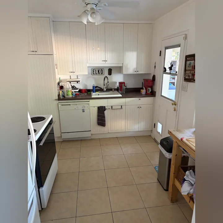 A tidy kitchen with white cabinets, a sink, stove, dishwasher, and a wooden table. Various kitchen items are visible on the countertop.