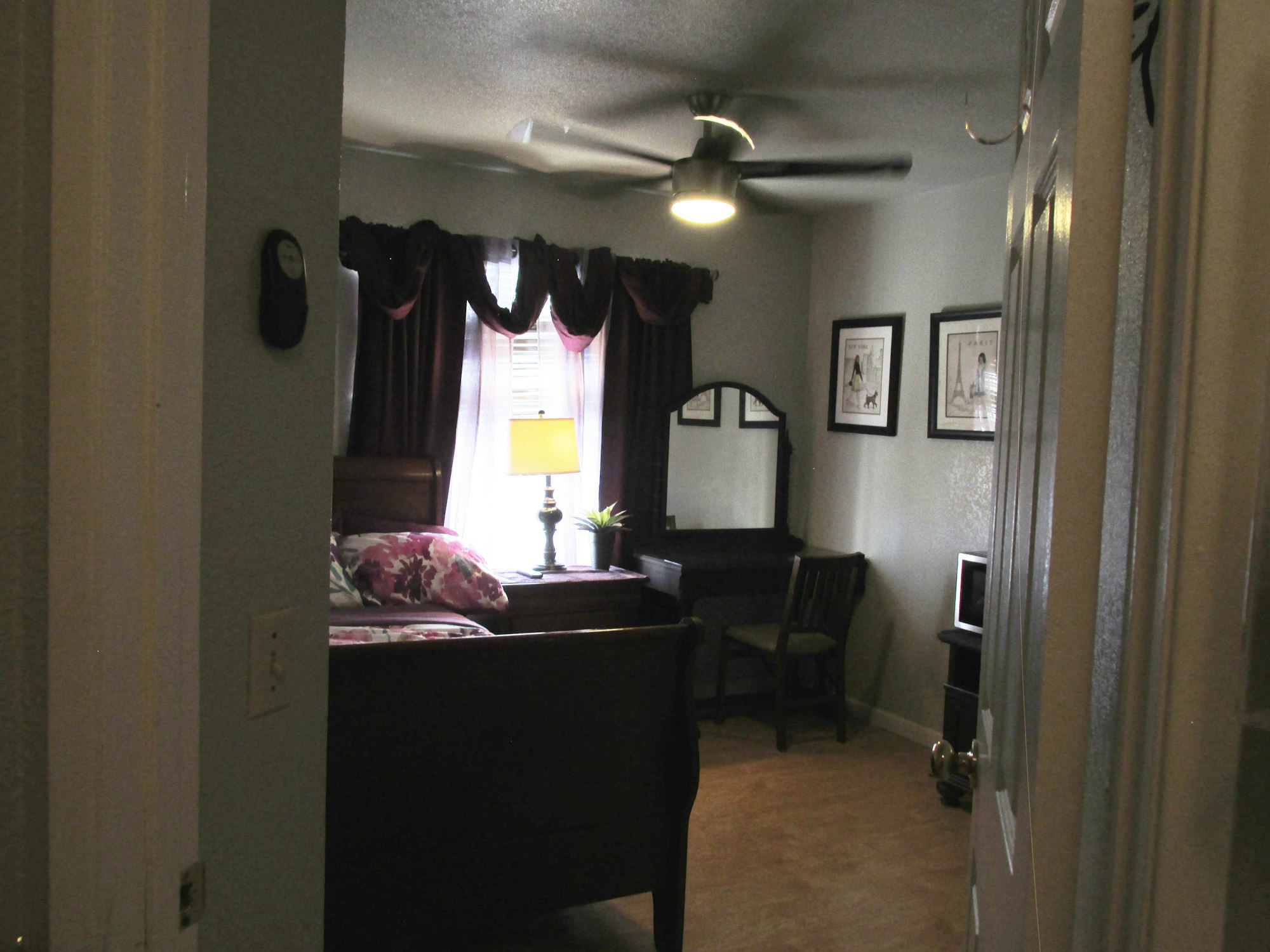 A bedroom with a bed, floral pillows, a nightstand with a lamp, a fan, a mirror on a vanity, curtains, and framed pictures on the wall.