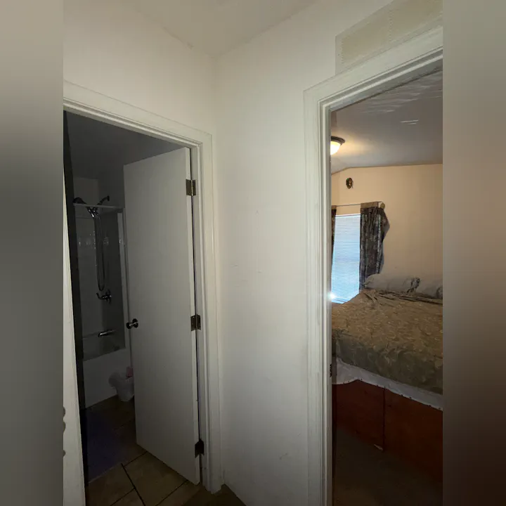 The image shows a view through a doorway, revealing a bedroom with a bed and a bathroom area with a shower.