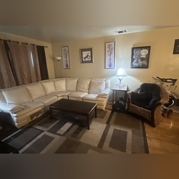 A cozy living room with a white sectional sofa, wicker chair, coffee table, lamp, wall art, curtains, and an exercise bike.