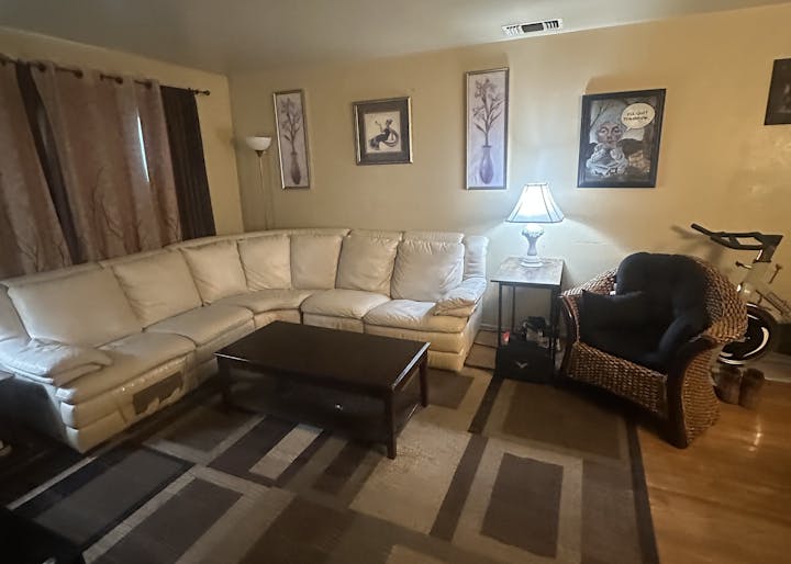 A cozy living room with a white sectional sofa, wicker chair, coffee table, lamp, wall art, curtains, and an exercise bike.