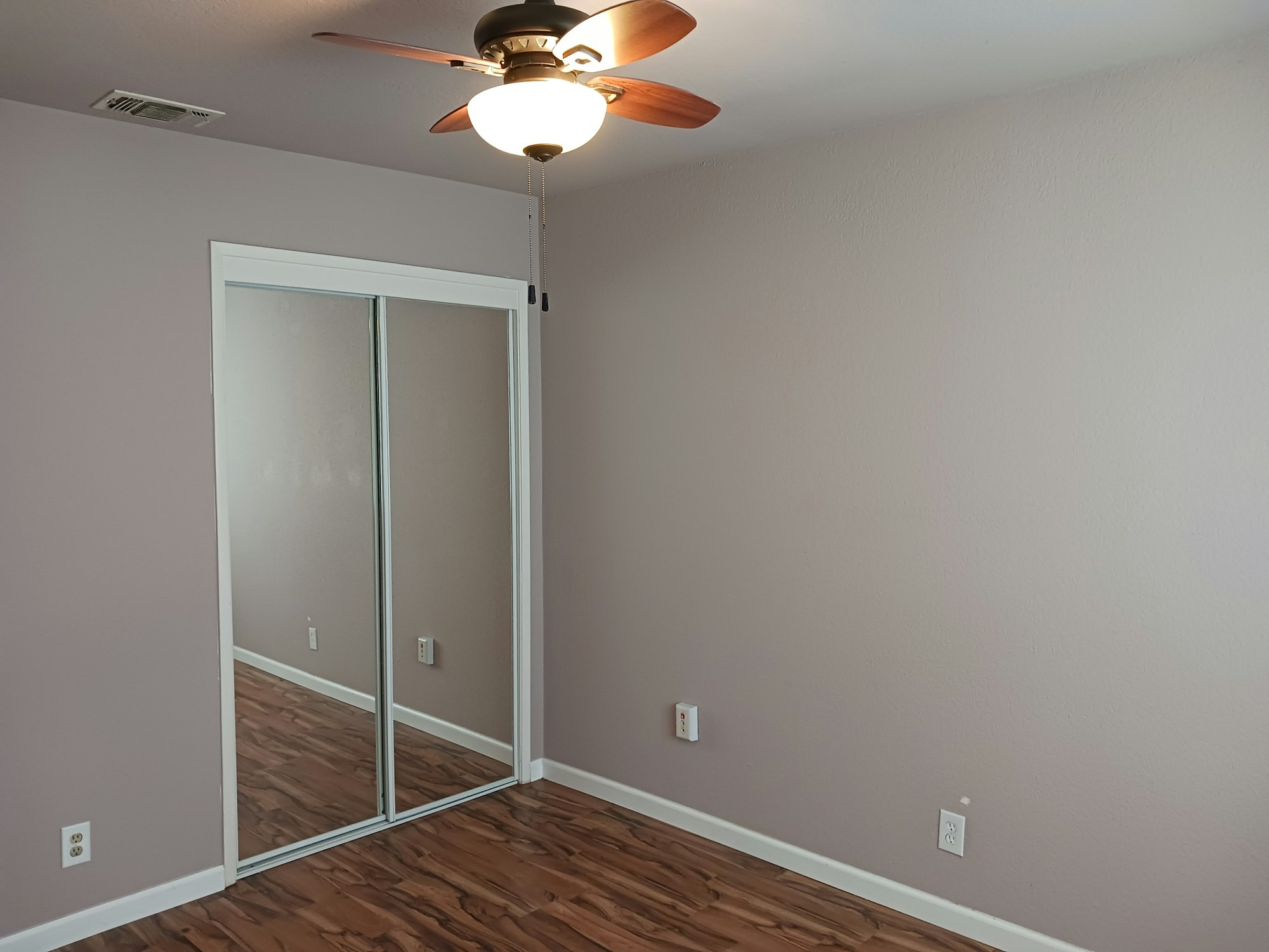 A simple room with a light ceiling fan, mirrored closet doors, a light purple wall, and laminate flooring.