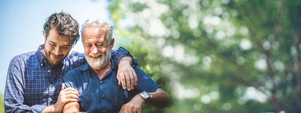 Two men, one younger and one older, smiling and embracing outdoors with blurred green foliage in the background.