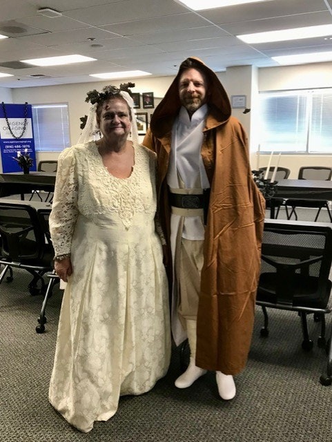 A woman in a vintage lace dress and a man in a Jedi costume pose together in a room with chairs and tables.
