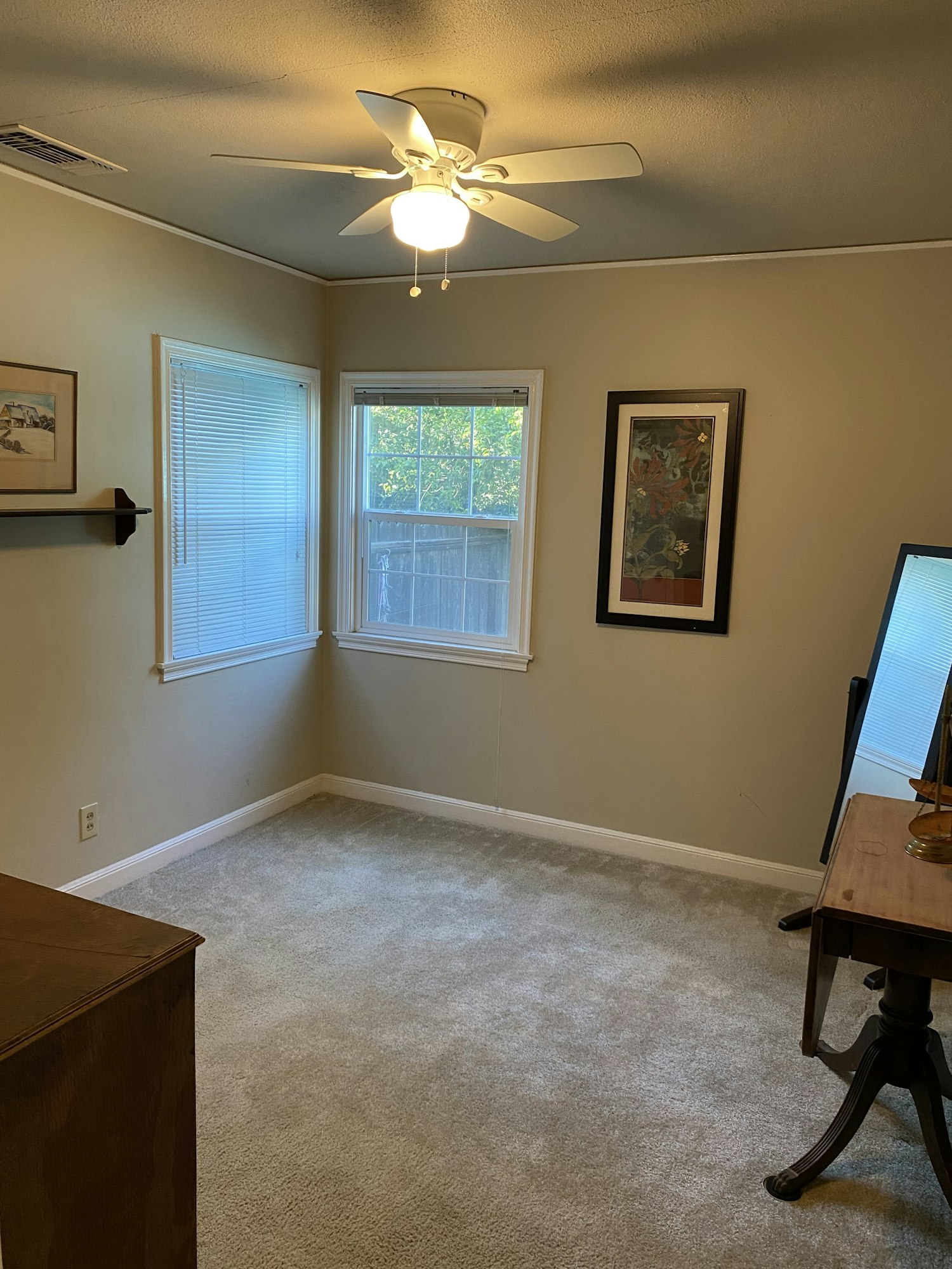 A room with carpet, a window, ceiling fan, paintings on the walls, and a small table with a lamp.